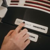Palec z pomalowanym na czarno paznokciem biegnie po piśmie brajlowskim. Napis: „orkiestron