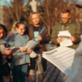 Grupa osób stoi na zewnątrz i wpatruje się w trzymane kartki. Na pierwszym planie trzymany wydruk z nagłówkiem: Aktor na deskach.