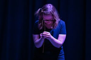 Kadr ze spektaklu w teatrze, jest ciemno. Młoda dziewczyna pochyla głowę, w wyciągniętych rękach trzyma kłos zboża.