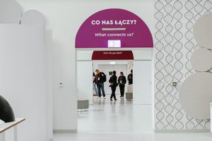 Białe, puste wnętrze. Szerokie przejścia prowadzą do kolejnych pomieszczeń. Nad pierwszym widać napisy: “Co nas łączy? What connects us?”, nad drugim tylko fragment: “How do you feel?”. W głębi ludzie w ruchu.
