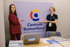 Dwie młode kobiety uśmiechają się do aparatu i pokazują logo Centrum Komunikacji dla Osób z Niepełnosprawnościami. Są elegancko ubrane, przed nimi na stoliku widać laptop i wiele kolorowych broszur.