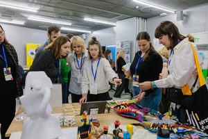Dynamiczna scena: kilka osób stoi wokół stolika, który jest pełen kolorowych przedmiotów z drewna i materiału. oglądają, dotykają, pani w centrum wskazuje na coś palcem.