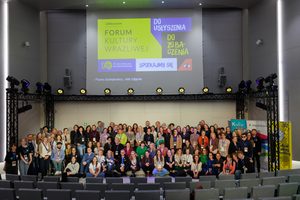 Bardzo wiele osób zebrało się na scenie, by zapozować do zdjęcia grupowego. Nad nimi wyświetla się baner Jubileuszowego Forum Kultury Wrażliwej z hasłami: do usłyszenia, do zobaczenia, spotkajmy się.