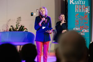 Elegancka kobieta na scenie trzyma teczkę i mówi do mikrofonu. W tle widać tłumaczkę języka migowego i baner Małopolskiego Instytutu Kultury.