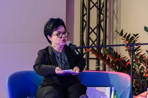 Elegancko ubrana niskorosła kobieta siedzi na kanapie na scenie. Trzyma plik kartek i mówi do mikrofonu.