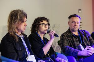 Trzy osoby siedzą w fotelach na scenie. Mężczyzna z długimi włosami, kobieta w okularach z kręconymi włosami oraz krótko obcięty mężczyzna w koszuli w kratę. Kobieta mówi coś do mikrofonu.