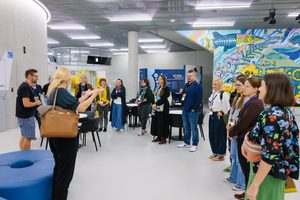 Grupa ludzi stoi w półokręgu, słuchając przewodnika. Tłumaczka przekłada jego słowa na język migowy. Są w nowoczesnym wnętrzu, jest dużo bieli, szkła i wielokolorowa ściana z namalowaną podmorską sceną.