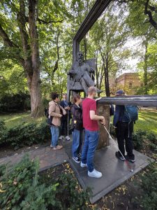 Grupka osób w otoczeniu zieleni. Stoją przy metalowym pomniku mężczyzny siedzącego w fotelu. Kilka osób trzyma białe laski i dotyka pomnika.