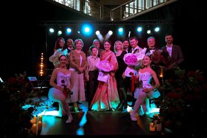 Grupa artystów pozuje na scenie, uśmiechają się do obiektywu. Niektórzy trzymają kwiaty.