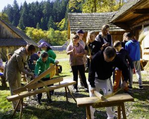 Zajęcia na świeżym powietrzu przed drewnianymi chatami. Dorośli i dzieci używają tradycyjnych przyrządów gospodarskich, ktoś trzyma wiązki słomy.