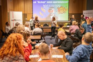 Wiele osób w sali. Prowadząca jest w ruchu, mówi do mikrofonu. Na ekranie teksty: "Jak Biszkopt został bohaterem" i "superhero" oraz czarno-białe zdjęcie chłopca z dodaną maską i błyskawicami. Wszyscy są aktywni, rozmawiają, ktoś zgłasza się z pytaniem.