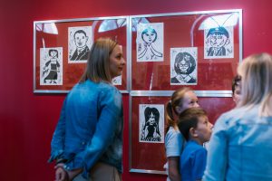 Kobieta i dzieci oglądają wystawę w galerii. Na tle ciemnoczerwonej ściany wiszą grafiki. Przedstawiają rożnych ludzi, jakby narysowanych przez dziecko.